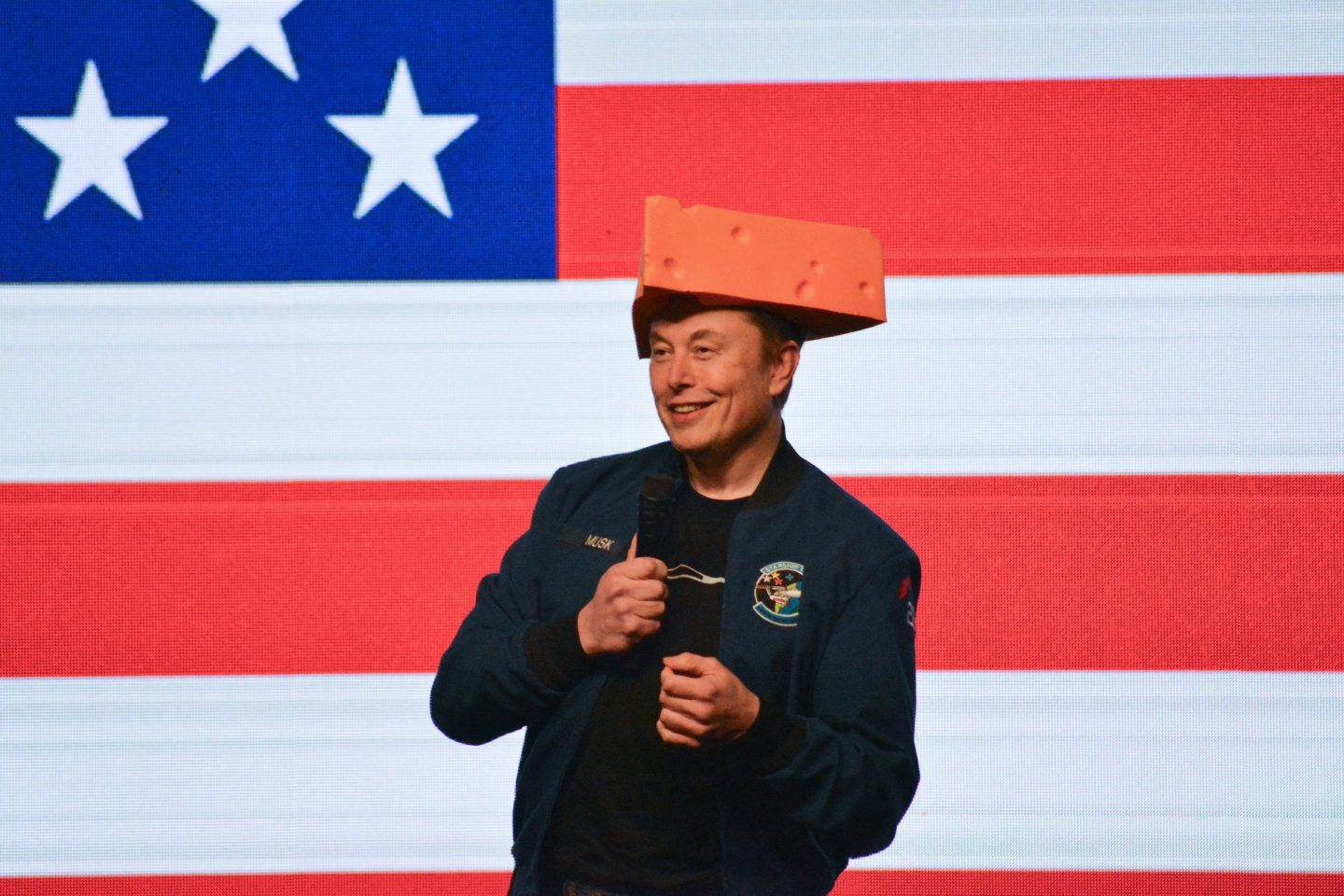 Elon Musk is wearing a cheese hat and standing in front of a giant American flag background.